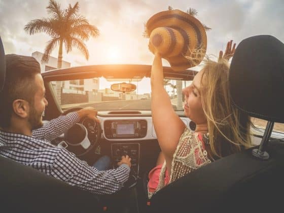 couple in car driving with woman's hands in the air, palm tree in background
