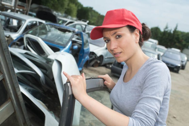 Woman in a red cap stands in a salvage yard holding a car door panel and pointing toward rows of wrecked vehicles, related to salvage title insurance California.