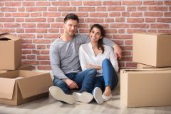 Young Smiling Couple Sitting Between Cardboard Boxes In Their New Home