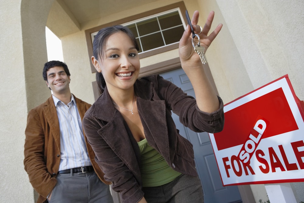 Young smiling Hispanic woman dangles keys to new home while husband looks on from behind