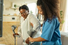 A young African American health workers helps and older female with crutches