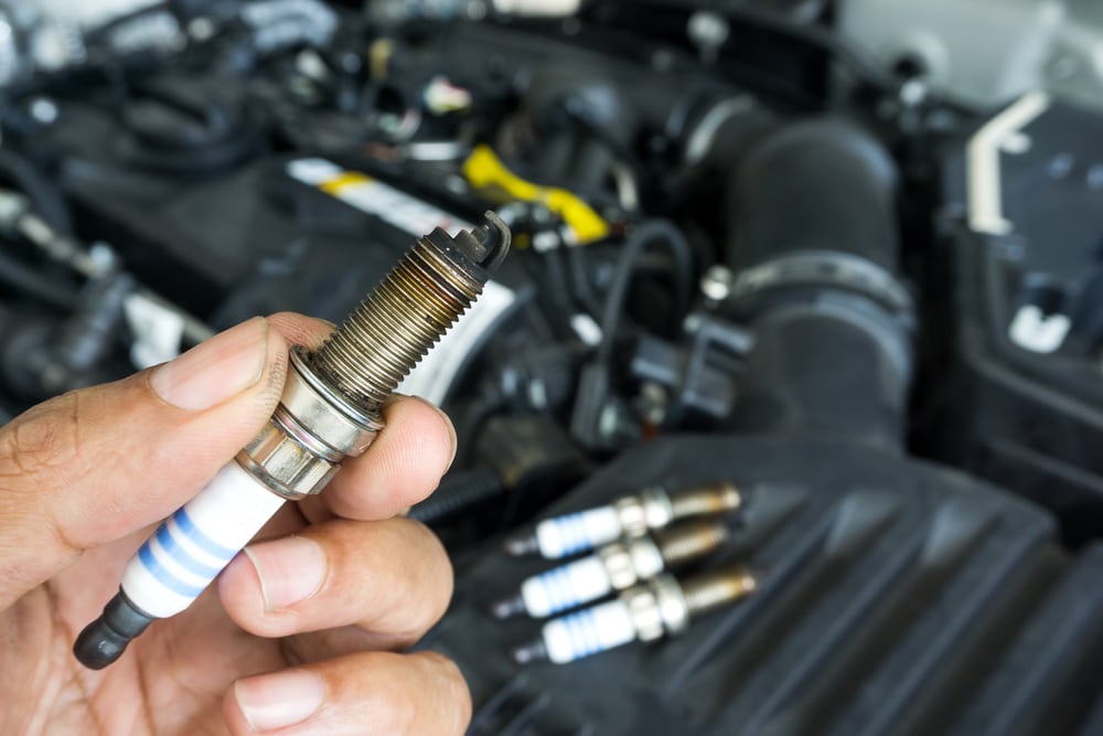 A hand holds a spark plug, blurred engine in the background