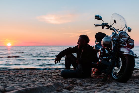 Motorcyclist sits on the beach next to his bike with sun setting.