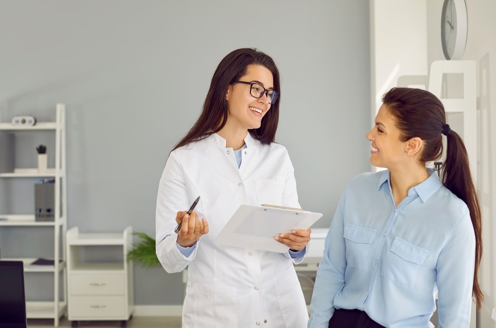 Friendly cheerful nurse or doctor at modern clinic talking to patient. Happy physician or gynecologist holding clipboard, smiling and giving medical consultation to young woman.