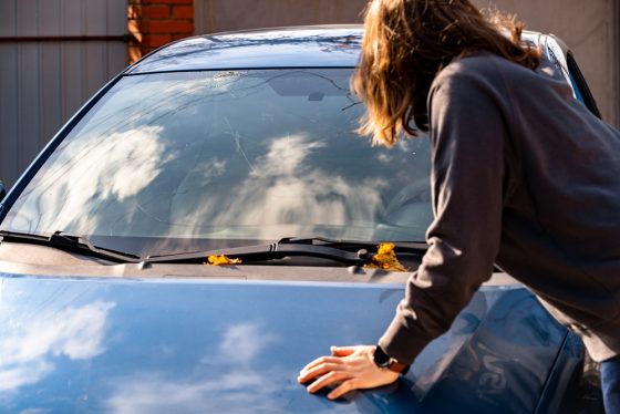 Woman with hands on hood of car looking at cracked windshield.