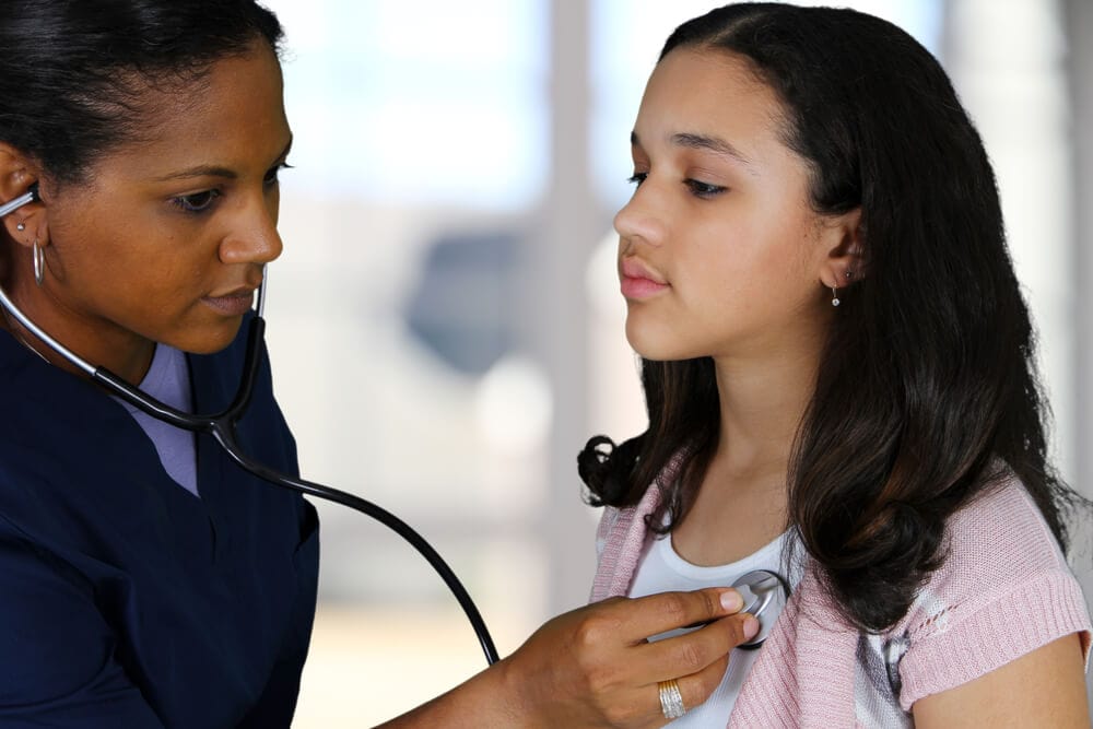 An African-American nurse listens to a young Hispanic girl's chest with a stethoscope.