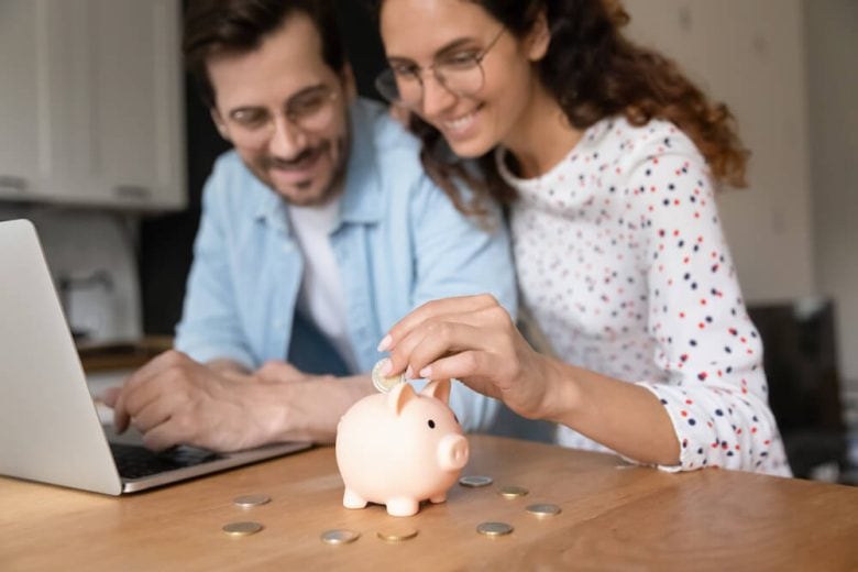 Happy young couple put savings in piggy bank while they shop around for cheap car insurance in California on laptop.