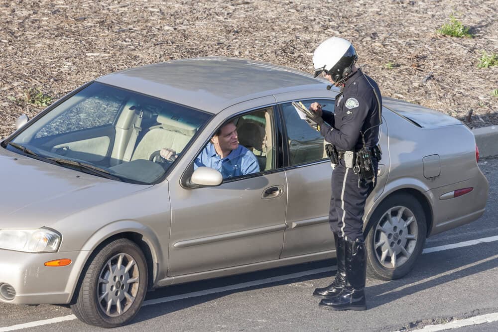 Police officer standing by a car and writing a speeding ticket while concerned driver looks on - cheap car insurance in California.