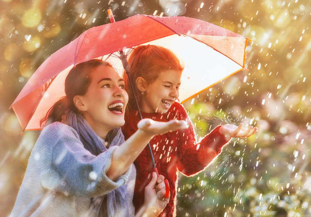 Mother and young daughter huddle under umbrella, laughing at the rain - cheap umbrella insurance in California.