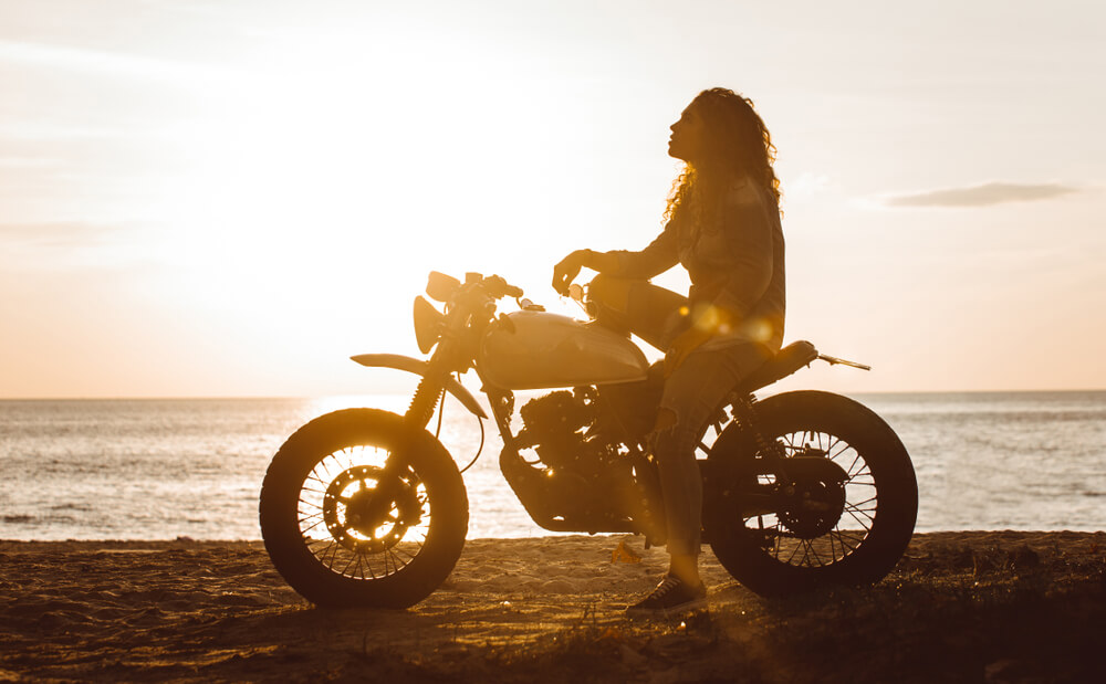 Young woman sits on her motorcycle parked in front of the ocean in California - cheap motorcycle insurance.