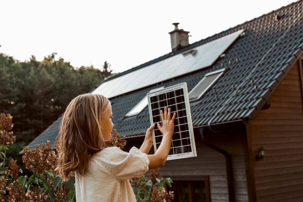 Little girl with model of solar panel, standing in the middle of meadow, house with solar panels behind. Concept of renewable resources, solar panels and home insurance in California.