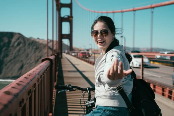 Girl backpacker, riding bike on Golden Gate Bridge, smiling inviting people to come to San Francisco. How to sign up for health insurance and car insurance when you move to California.
