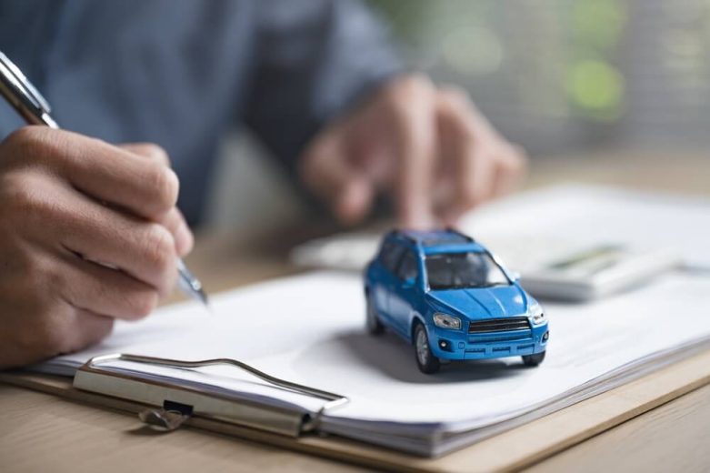 A small blue toy car is on top of insurance documents with a blurred man’s arms in the background holding a pen, concept of the cost of insurance after a lapse in coverage.