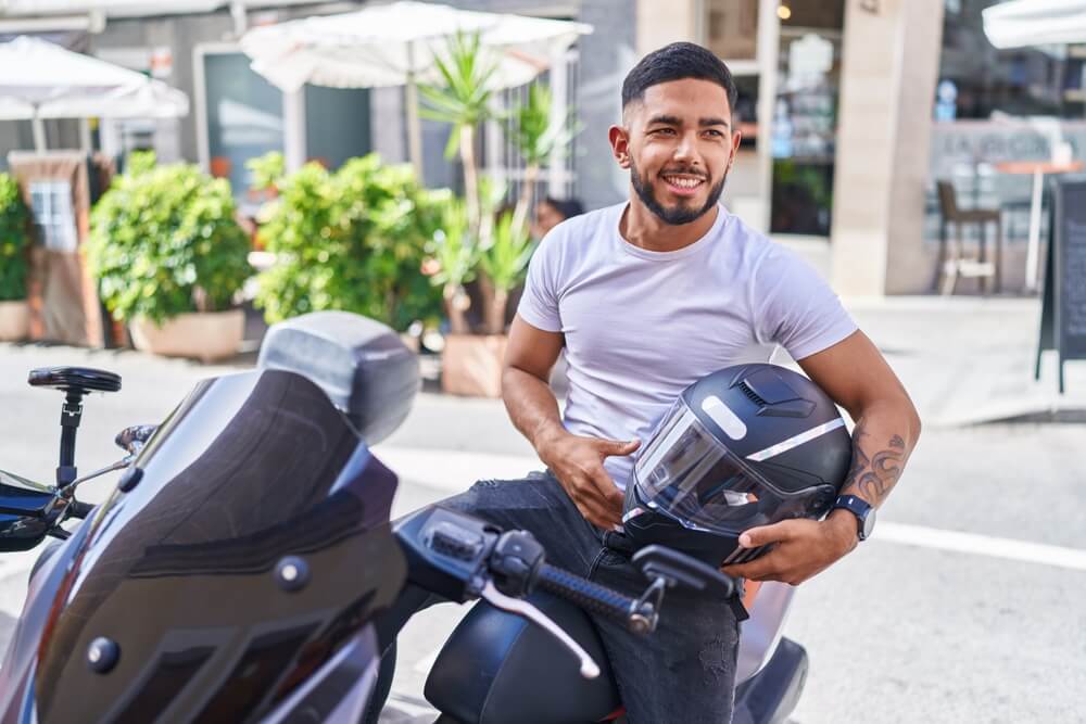 Latino man on a motorcycle holding his helmet and smiling after receiving insurance tips for motorcycle owners in California from Cost-U-Less.