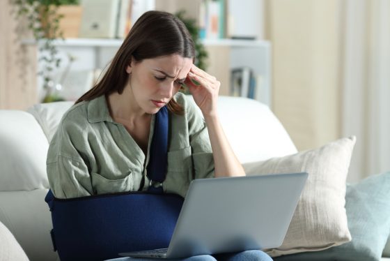Worried woman with injuries reading bad news that her health insurance claim is denied in California on laptop sitting on a couch in the living room at home.
