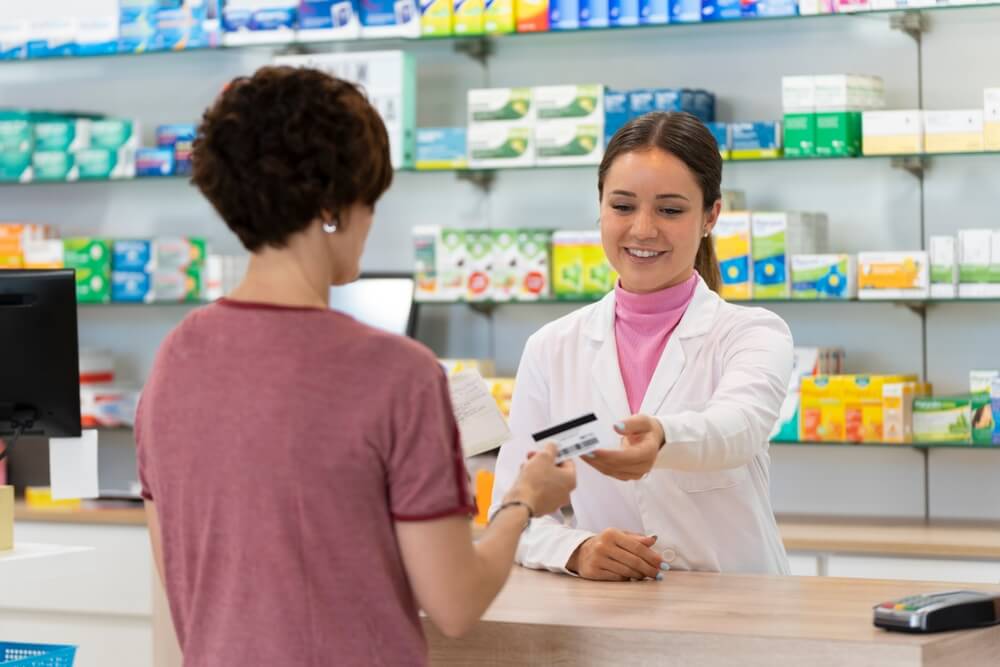 Pharmacist handing a customer her prescription and health card at the counter, showing how health insurance for part time workers in california helps with medication costs.