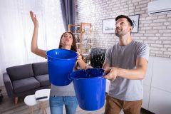 Couple holds buckets under a ceiling leak as water pours into their apartment, a real-life example of renters insurance water damage in California.