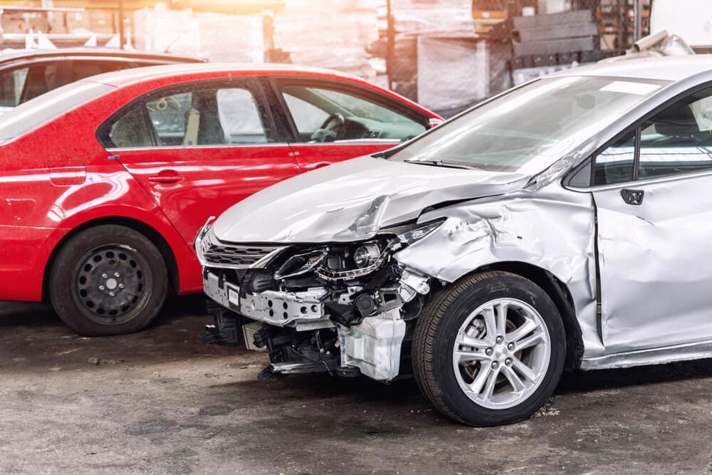 Badly damaged silver car with a crushed front end sits in a lot next to a red sedan, illustrating concerns about salvage title insurance California.