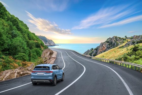 Scenic coastal highway curves along green hills toward the ocean as a blue SUV drives ahead, representing long-distance travel and car insurance for cross-state commuters in California.
