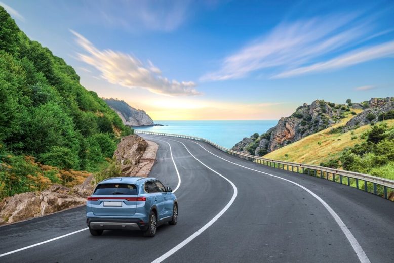 Scenic coastal highway curves along green hills toward the ocean as a blue SUV drives ahead, representing long-distance travel and car insurance for cross-state commuters in California.