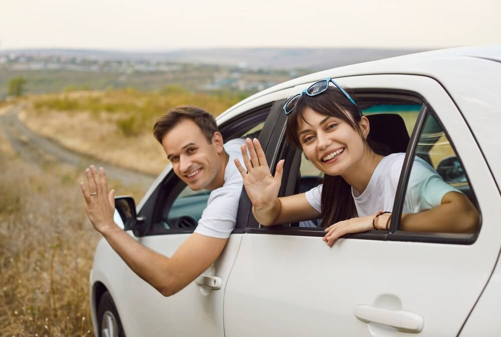 Smiling couple waves from the windows of a white car on an open road, representing travel routines and car insurance for cross-state commuters in California.