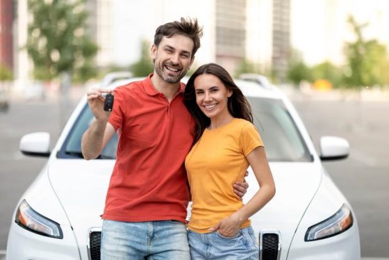 Smiling couple standing in front of a white car while holding up the keys, illustrating how to insure a car not in your name.