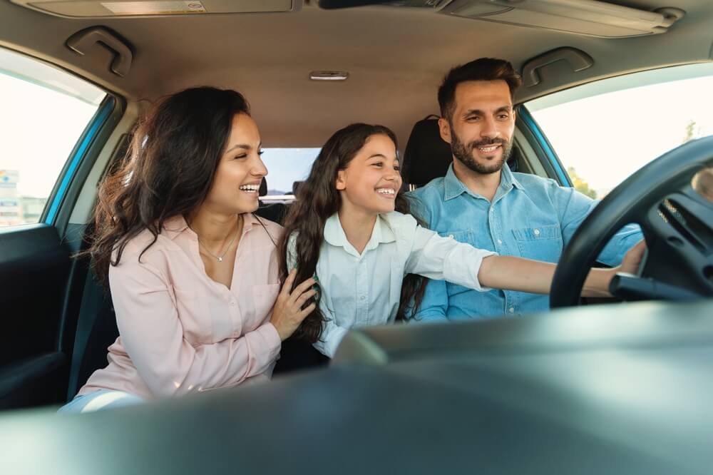 Family smiling inside a car as a child reaches toward the steering wheel, illustrating how to insure a car not in your name.