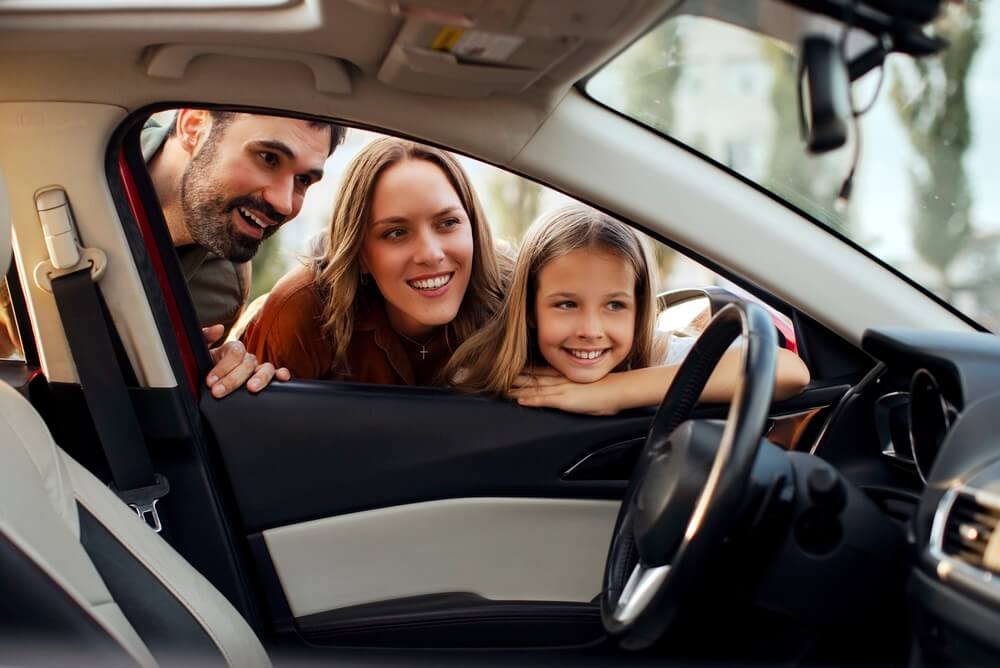 A smiling family leans into a car through the open door while discussing paperwork about how to insure a car not in your name.