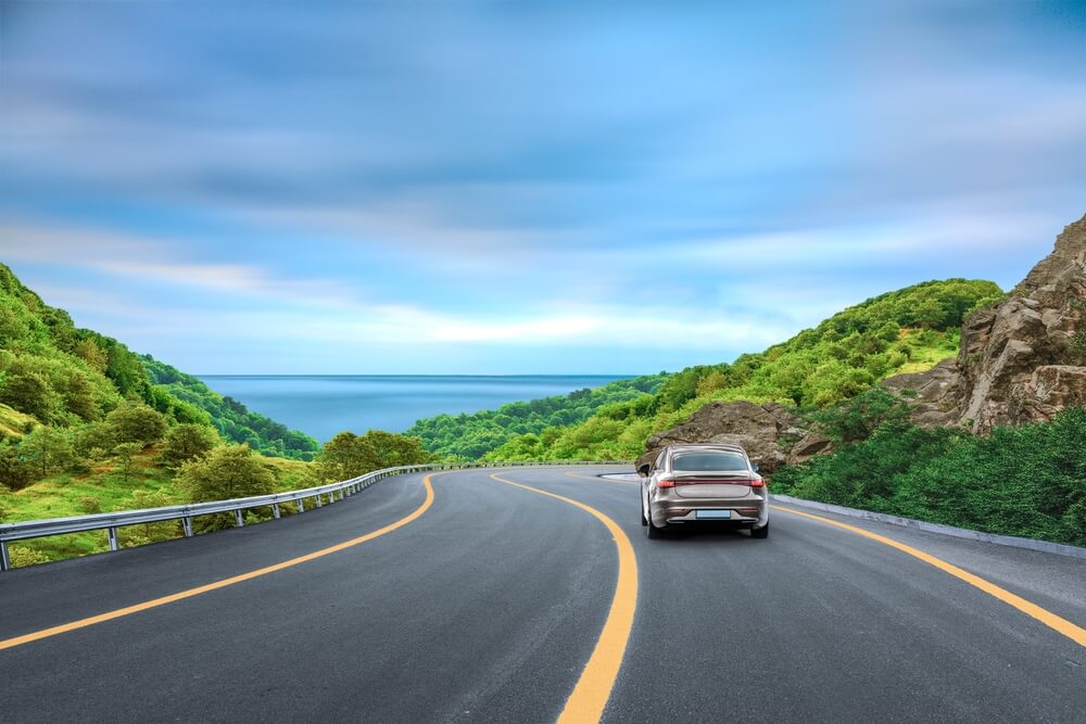A car driving along a winding coastal highway under a bright blue sky, illustrating new car insurance rules in california.