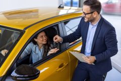 A car salesperson in a showroom hands a set of car keys to a smiling customer seated in a bright yellow car. The salesperson is holding a clipboard, and the scene suggests the purchase or rental of a vehicle, relating to auto insurance or rental car coverage.