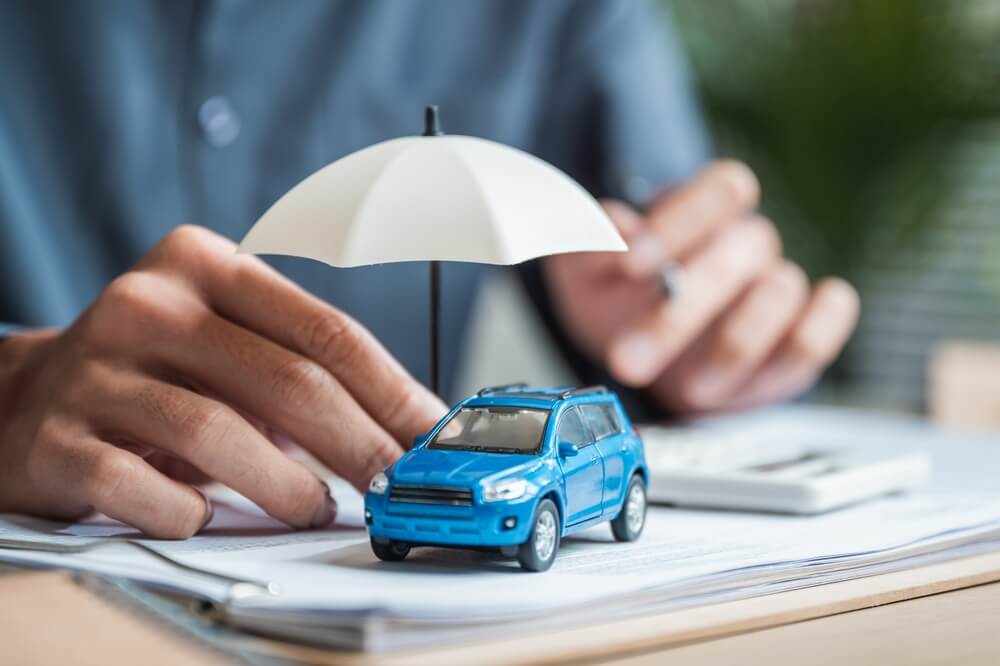 A miniature blue SUV sits beneath a white umbrella on top of paperwork while a person’s hands are positioned nearby. The image symbolizes auto insurance protection, coverage, and financial security for vehicles, including rental car coverage.