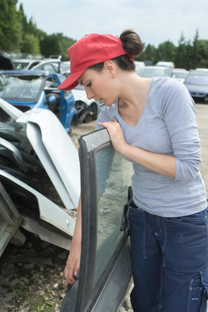 A person in a red cap examines a removed car door in a salvage yard filled with salvaged vehicles and auto parts.