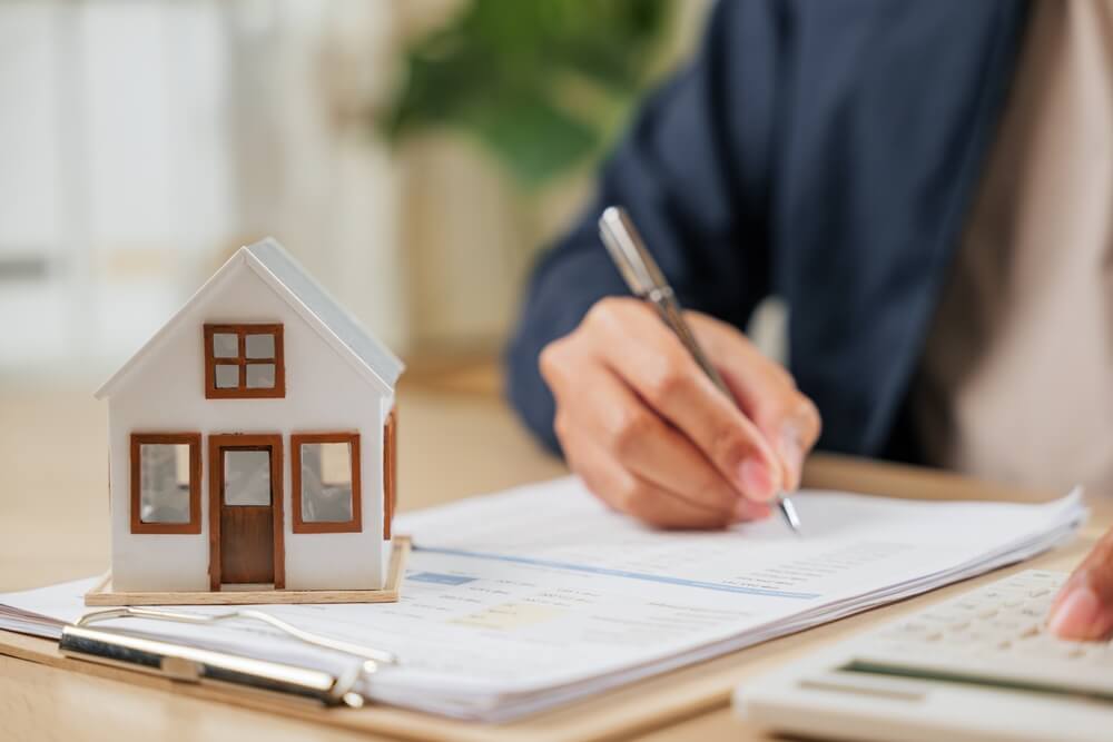 Person signing paperwork beside a model house, illustrating house insurance considerations for a vacant California home.