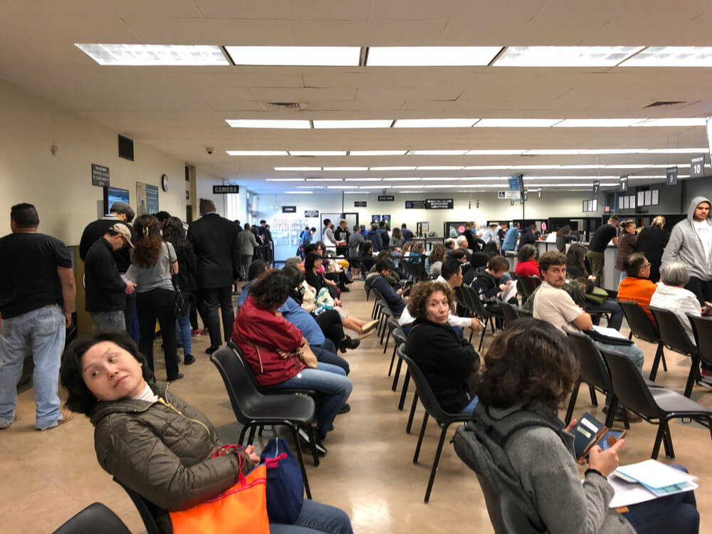 People wait in a busy California DMV office with service counters in the background, showing why planning ahead matters for how to get in and out of the DMV fast in California.