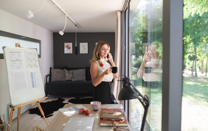 Woman takes a phone call inside a modern backyard office with a desk, laptop, and presentation board, connecting the workspace to home insurance if you build a backyard office or studio in california.