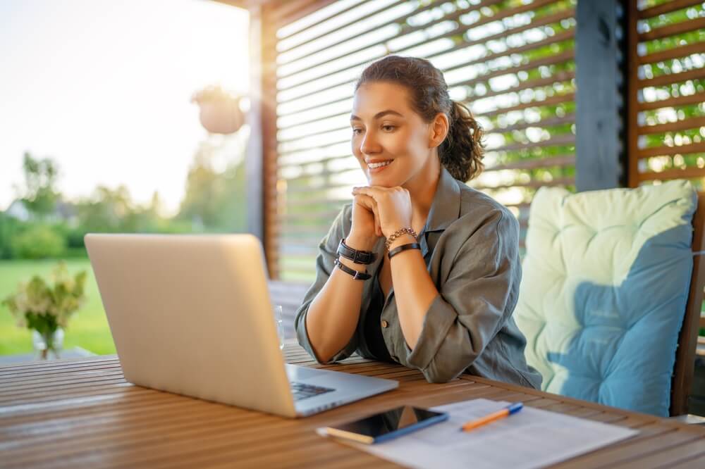Woman working on a laptop from an outdoor backyard workspace, connecting remote work setups with home insurance if you build a backyard office or studio in california.