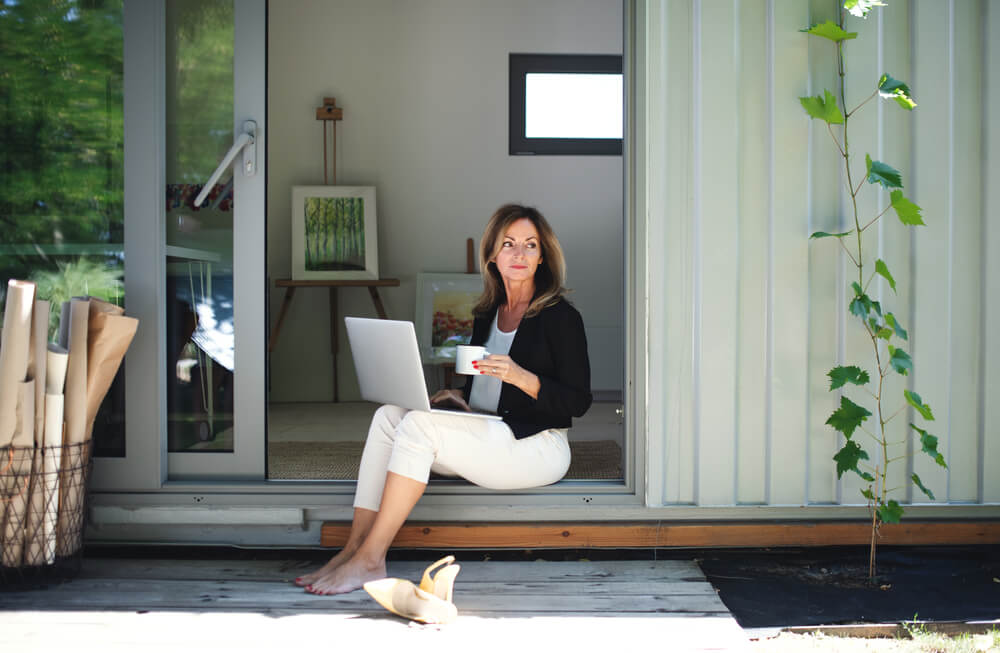 Woman sits at the entrance of a backyard studio with a laptop and coffee, connecting remote workspaces with home insurance if you build a backyard office or studio in california.