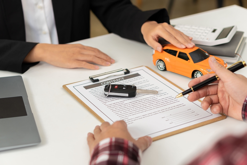 Customer reviews and signs paperwork for a branded car title transfer while an agent presents a model vehicle and car keys on a contract desk.