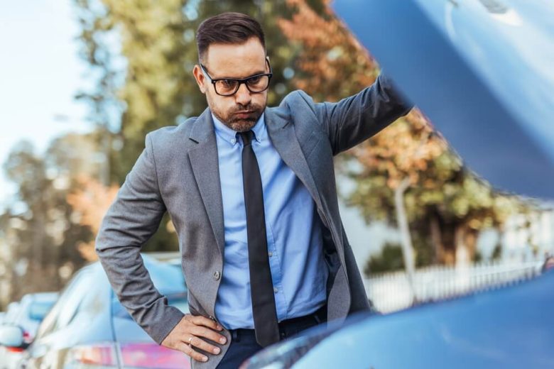 Businessman inspecting an open car hood after a breakdown, illustrating mechanical failures and car insurance coverage.