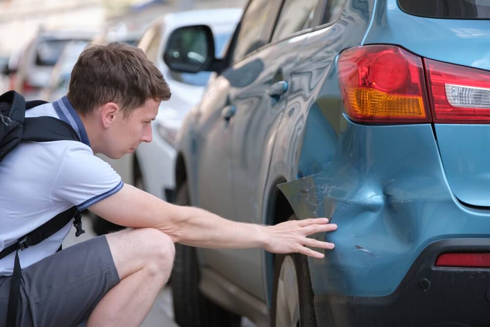 Driver inspecting a dented rear bumper on a parked car, showing the aftermath of a parked car hit and run in California.
