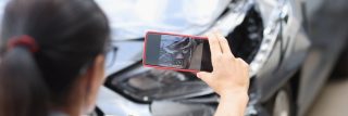Driver taking a photo of front-end damage with a phone, documenting evidence after a parked car hit and run in California.