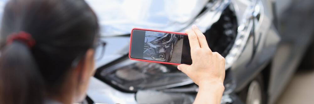 Driver taking a photo of front-end damage with a phone, documenting evidence after a parked car hit and run in California.