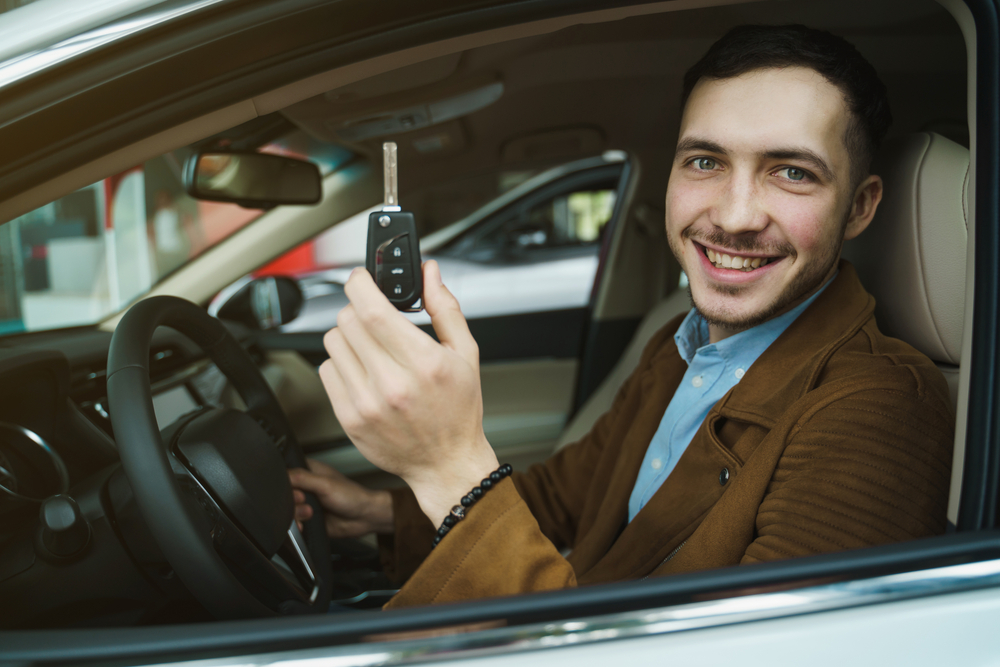 Smiling driver sits behind the wheel holding up a car key, illustrating a choice between high and low auto insurance deductibles.