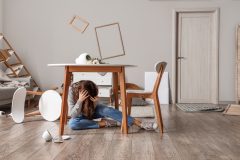 Persona sentada en el suelo bajo una mesa en una vivienda con objetos caídos tras un terremoto, representando los daños y la protección que ofrece el seguro para viviendas en zonas sísmicas de California
