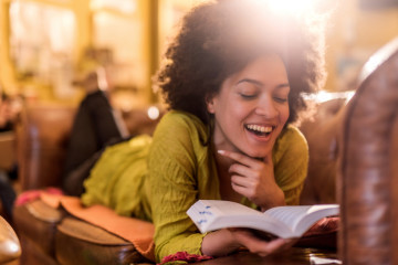 Cheerful African American woman relaxing on sofa at home and reading a book.