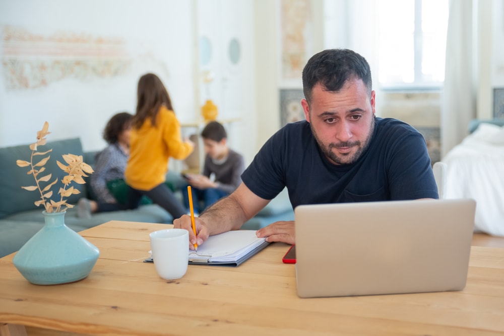 man looking at laptop and taking notes with family on the background