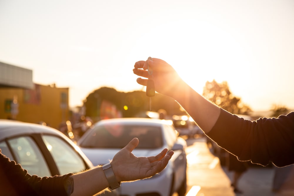 one hand handing car keys to another hand with vehicles in background