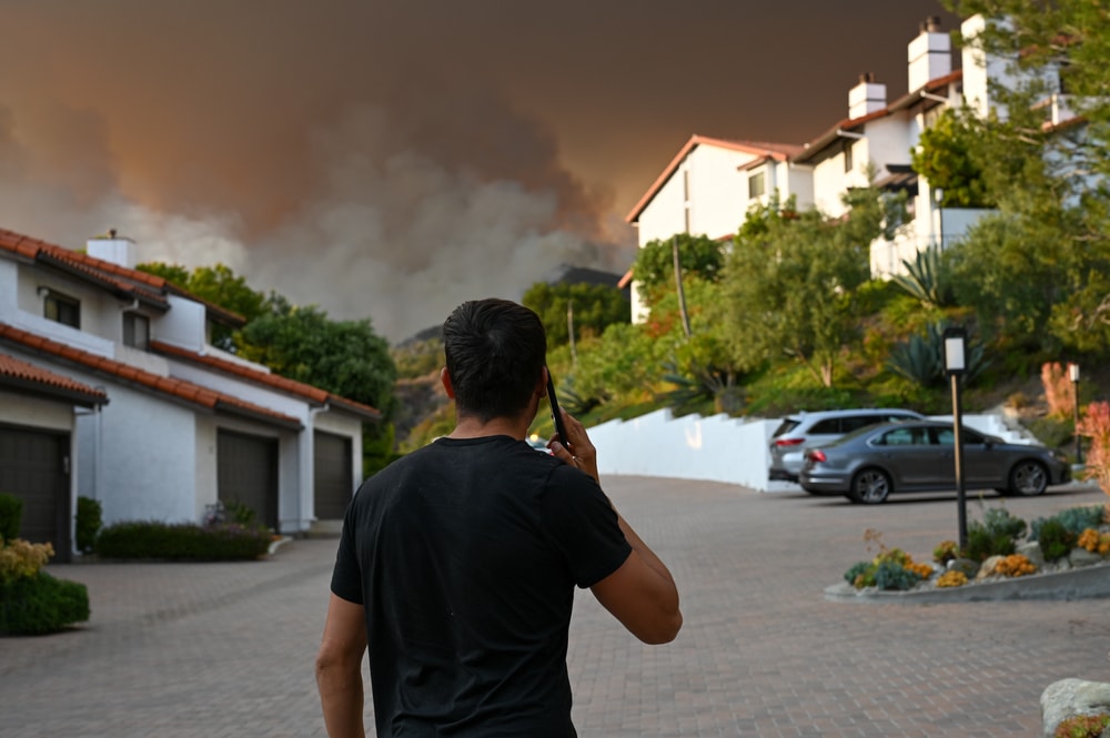 man standing on cell phone by homes watching california wildfire in distance