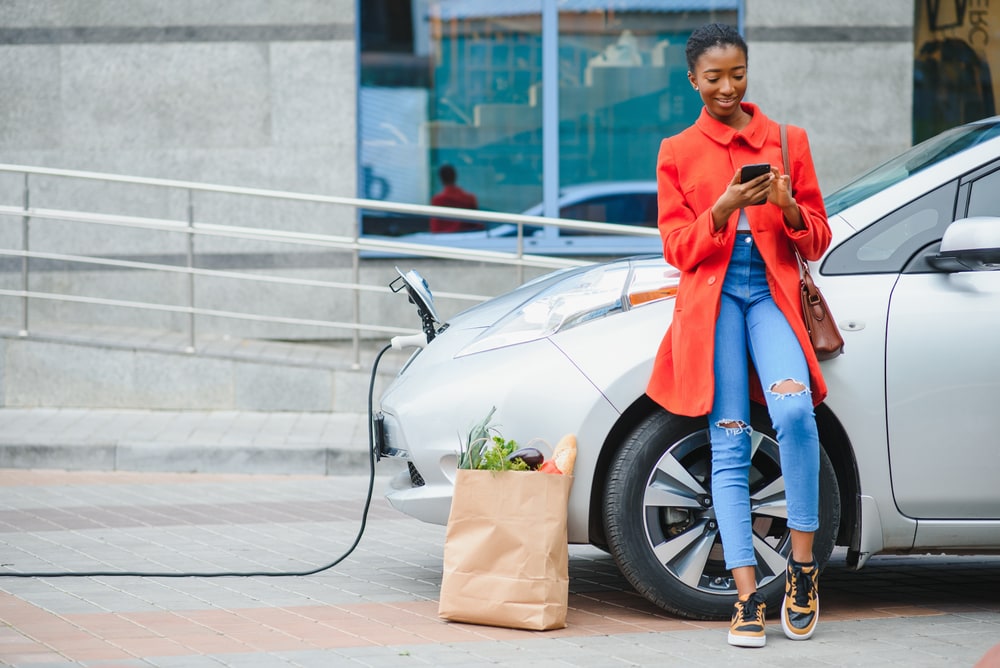African American woman looks at her phone while her electric car is charging