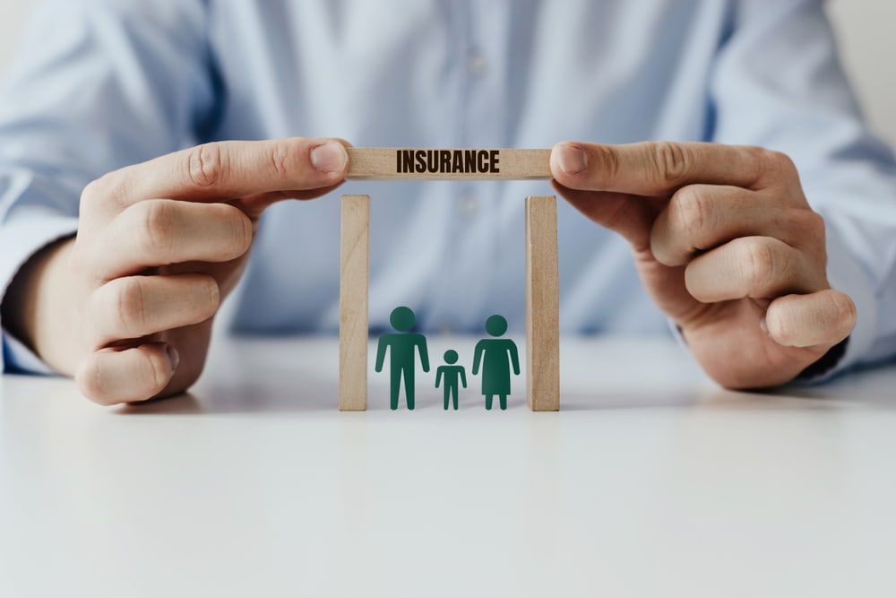 The businessman's hand covers the wooden house with a block with the inscription INSURANCE. Family at home with a roof with inscription insurance. Family insurance concept.